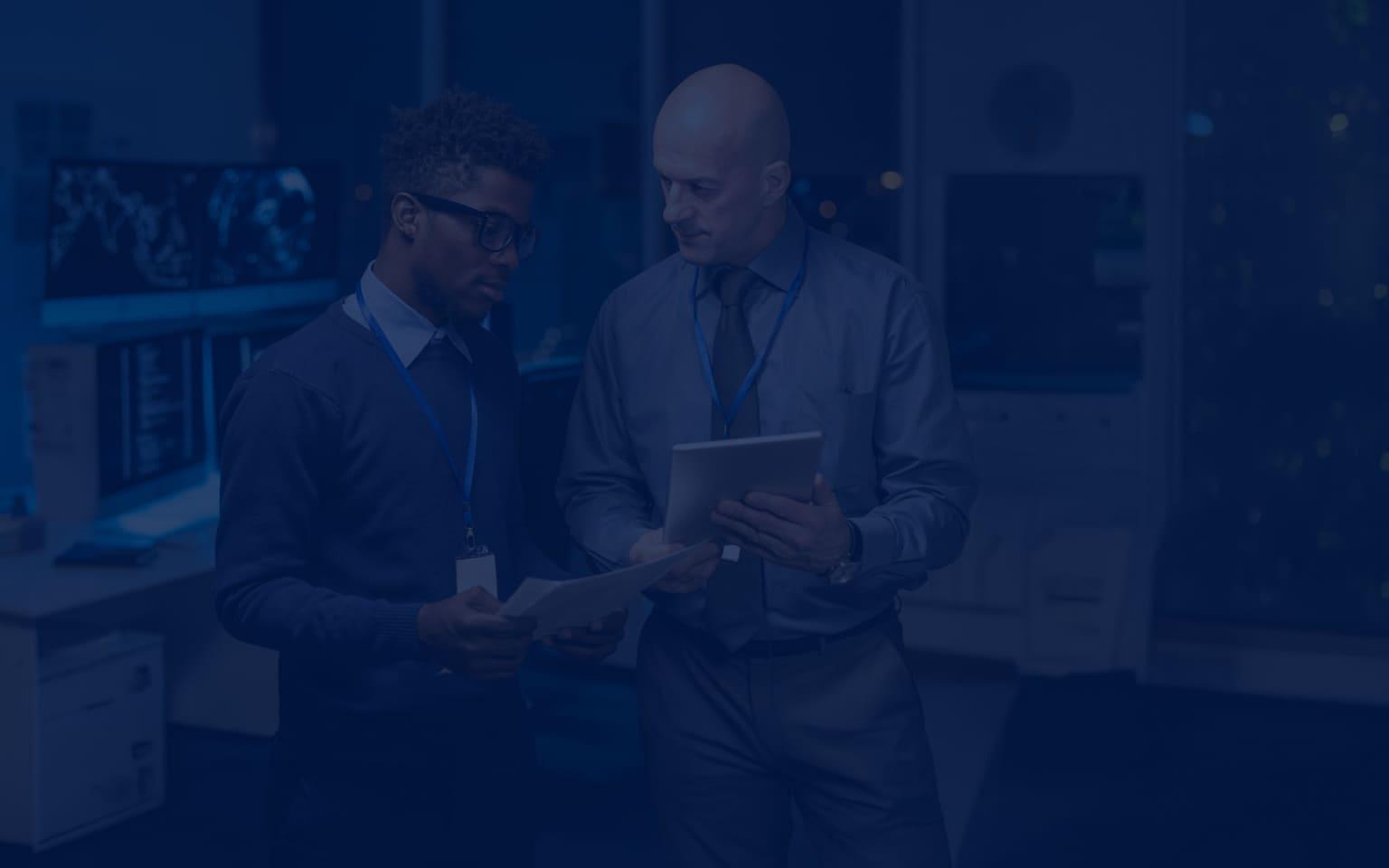 Zwei Männer in Anzügen stehen nebeneinander mit einem Tablet in der Hand, als Sinnbild für die Zusammenarbeit beim Bedrohungsmanagement.