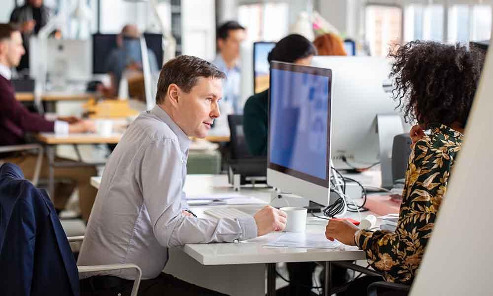 Automated workflows A man and woman seated at a desk in an office, engaged in discussions about automated ticket assignment and risk management.